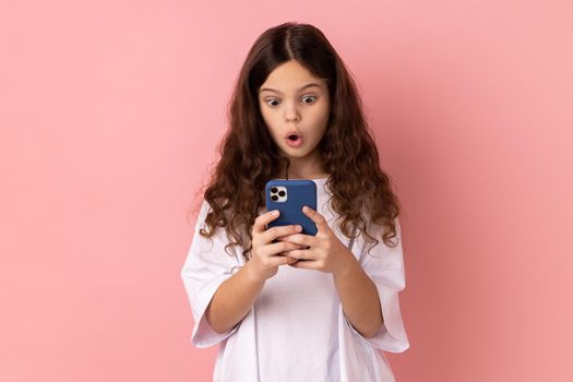 Mobile connection. Portrait of shocked astonished little girl wearing white T-shirt reading shocking post on social network using cell phone. Indoor studio shot isolated on pink background.