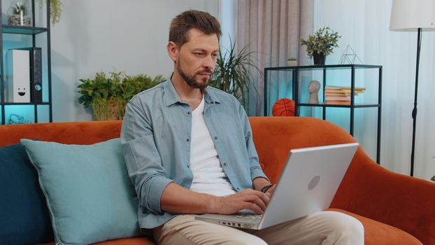 Portrait of man sitting on couch closing laptop pc after finishing work in living room at home sofa. Young guy freelancer working online, remote job. E-learning, browsing internet on notebook computer