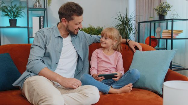 Happy family father or nanny and child kid daughter laugh use smartphone app look at screen play online game, daddy with child take selfie watch cartoons make video call on mobile sit on sofa at home