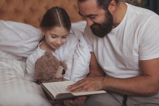 Cute little girl hugging her teddy bear, lying in bed while her father reading her a book before sleep. Handsome mature bearded man enjoying reading fairytales to his lovely daughter