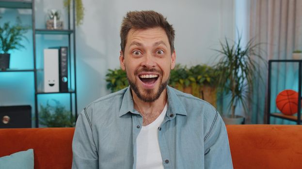 Oh my God, Wow. Handsome adult man surprised looking at camera with big eyes, shocked by sudden victory, good win news, celebrating at home. Excited amazed young guy indoors in living room on couch