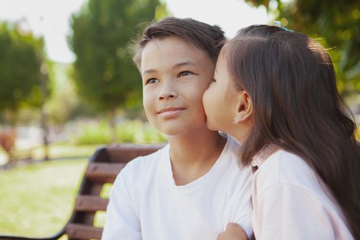 Close up of a young Asian boy smiling, looking away, his little sister kissing him on the cheek, copy space. Cute little girl kissing her brother on the cheek