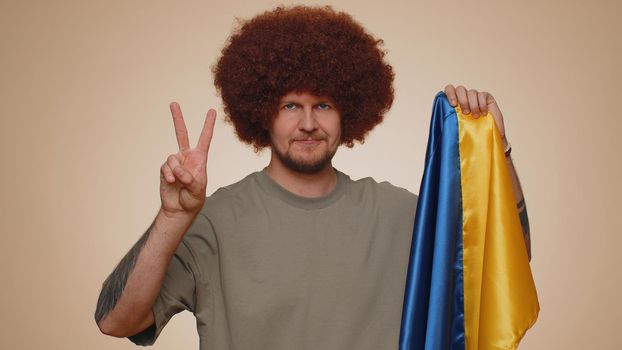 Man holding Ukraine national flag, showing victory sign against war, hoping for success and win, doing peace gesture, smiling with kind optimistic expression. Young guy boy on beige studio background