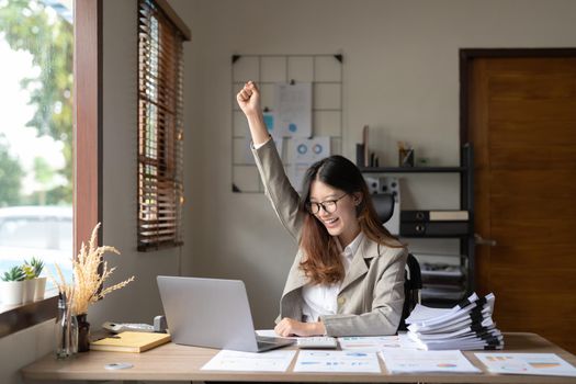 Successful young asian business woman achieving goals excited raised hands rejoicing with laptop in the office. New startup project concept