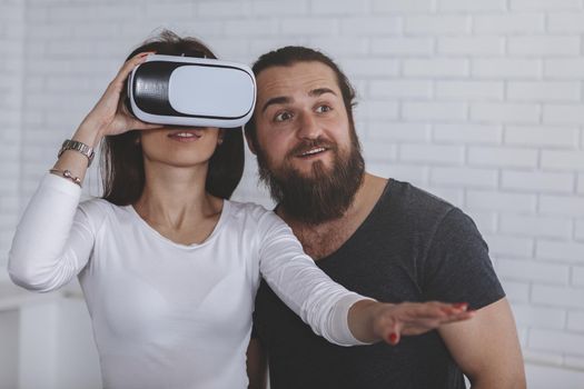 Cheerful bearded man and his girlfriend trying virtual reality glasses for the first time, copy space. Woman wearing 3d vr goggles, her biyfriend standing by. Technology, imagination concept