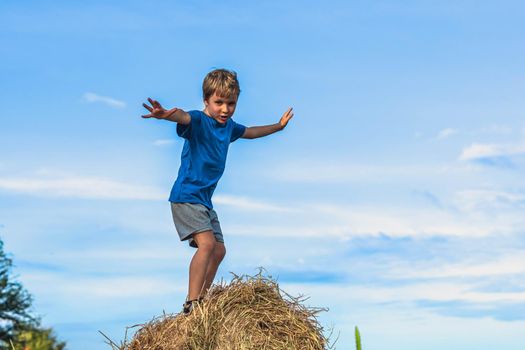 Boy smile play dance grimace show off blue t-shirt stand on haystack bales of dry grass, clear sky sunny day. Balance training. Concept happy childhood, children outdoors, clean air close to nature.