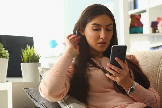 Serious beautiful woman sits on sofa and looks at smartphone screen. Smartphone applications reading news chatting