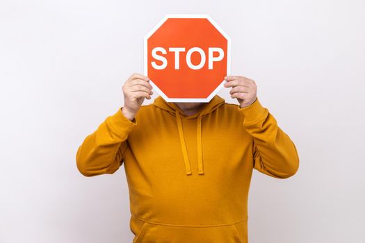 Attention, prohibited way. Unknown person hiding face behind Stop symbol, red traffic sign warning of restricted access, wearing urban style hoodie. Indoor studio shot isolated on white background.