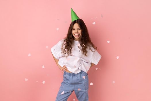 Little girl wearing white T-shirt and party cone standing under falling confetti, looking at camera smiling, celebrating birthday holiday, festive mood. Indoor studio shot isolated on pink background.