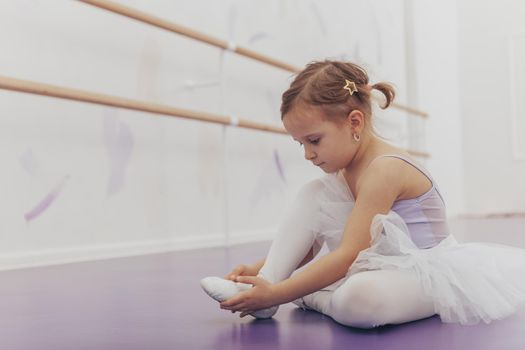 Adorable little girl putting on her ballet dancing shoes, sitting on the floor at dance studio. Lovely little ballerina preparing for exercising at ballet school, copy space. Achievement, dedication concept