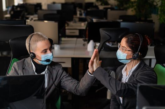 Two business women in masks are giving a high five while sitting at one desk in the office.