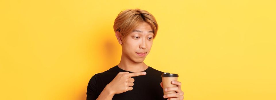 Close-up of impressed handsome asian guy pointing finger at delicious coffee, standing amazed over yellow background.