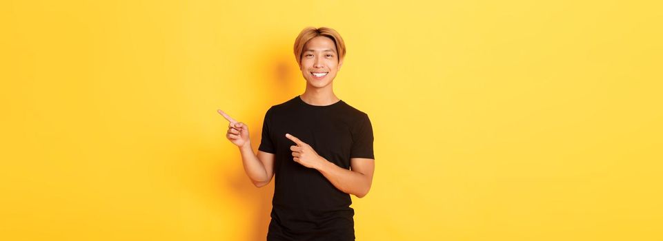 Portrait of satisfied smiling blond guy, asian student pointing fingers right, showing way, standing yellow background.