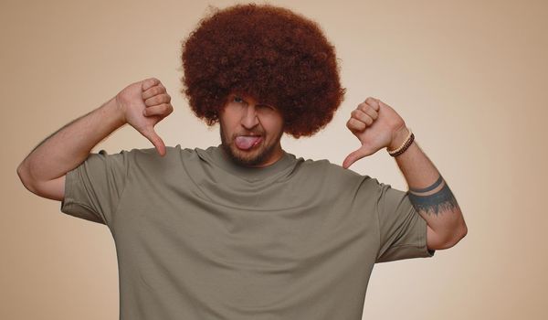 Dislike. Upset unhappy handsome man in t-shirt showing thumbs down sign gesture, expressing discontent, disapproval, dissatisfied, dislike. Young adult guy. Indoor studio shot on beige background