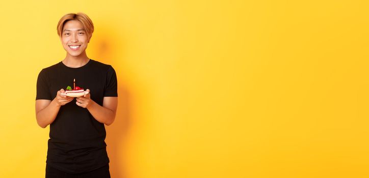 Handsome happy asian blond guy, smiling pleased as celebrating birthday, holding b-day cake, standing over yellow background.