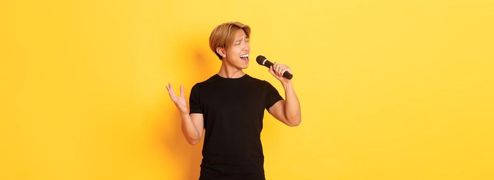 Carefree handsome asian guy, korean singer singing into microphone with passion, standing yellow background.