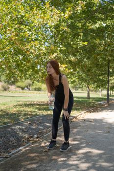 Full body young sportswoman with bottle of water bending forward and looking away while taking break in running session on sunny summer day in park