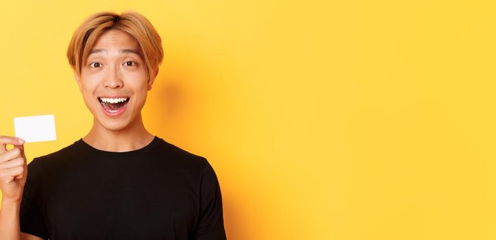 Close-up of excited surprised handsome asian guy, smiling amazed and showing credit card, standing yellow background.