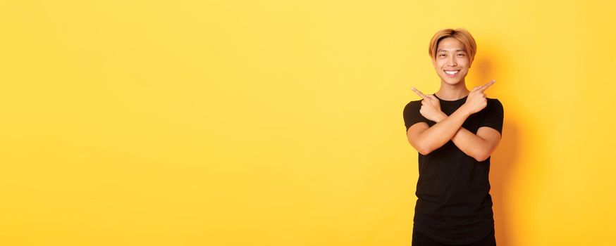 Portrait of handsome determined blond asian guy, student pointing fingers sideways, showing two variants, standing yellow background.