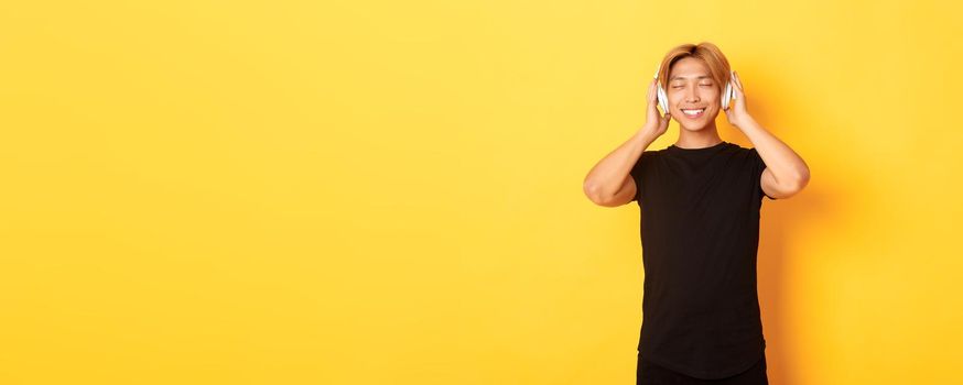 Relaxed and pleased handsome asian guy close eyes from satisfaction while listening music in wireless headphones, standing yellow background.