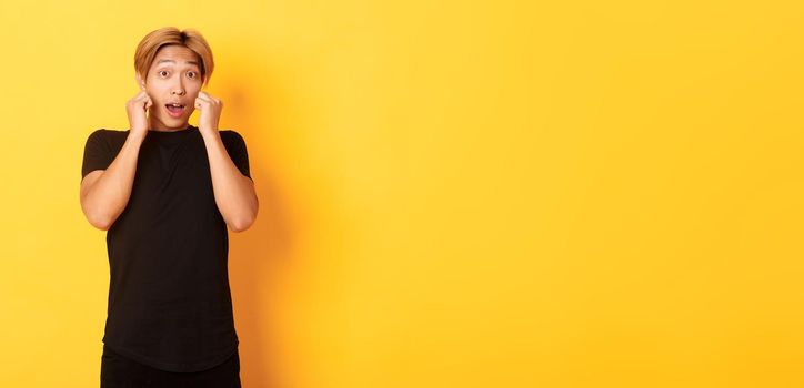 Excited handsome blond guy in black clothes, looking tempted and standing over yellow background.