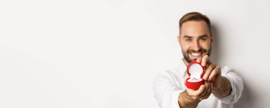 Close-up of handsome man asking to marry him, focus on box with wedding ring, concept of proposal and relationship, white background.