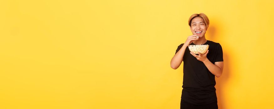 Carefree happy asian guy with blond hair, watching comedy, laughing and eating popcorn, standing yellow background.