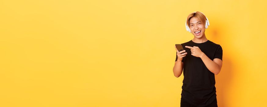 Joyful satisfied asian handsome guy, listening music or good podcast in headphones, pointing finger at smartphone with pleased smile, yellow background.