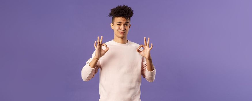 Portrait of skeptical young hispanic man rate something normal quality, show okay sign and grimacing judgemental, saying its not bad but average, standing purple background.