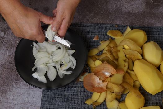 woman cutting onion on a black plate next to it peeled potatoes and peelings