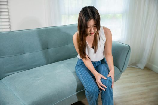 Asian young woman sitting on sofa and feeling suffering knee pain in living room at home, female massage her knee leg pain with painful face, Health bone problems self physiotherapy