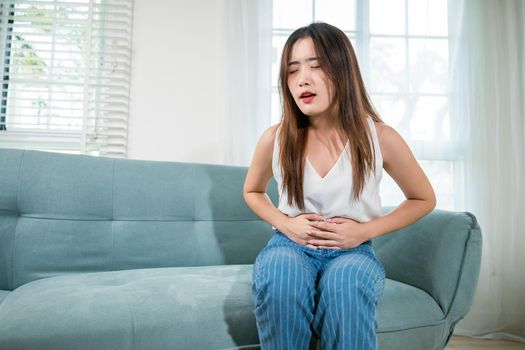 Asian young woman unhappy hands holding on stomach suffering from abdominal pain on sofa at home in living room, Sad female close eyes she painful stomachache, medical care and health concept