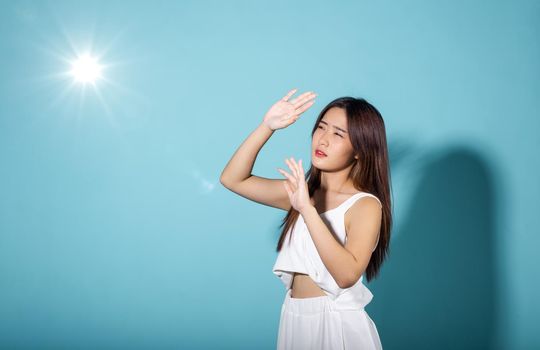 Asian young woman hand up cover face protect sun light and UV smile in studio shot isolated on blue background, female raising arm and hand in protection and block sunshine, sunscreen cream advertise