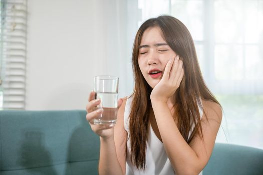 Beautiful female toothache and dental problems touching cheek feeling pain after drinking cold water, Asian young woman with sensitive teeth holding glass of cold water at home in living room, pain