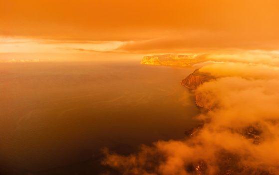 A red burning sunset with the silhouette of a cliff over the sea. Abstract nature summer or spring ocean sea background. Small waves on golden warm water surface with bokeh lights from sun.