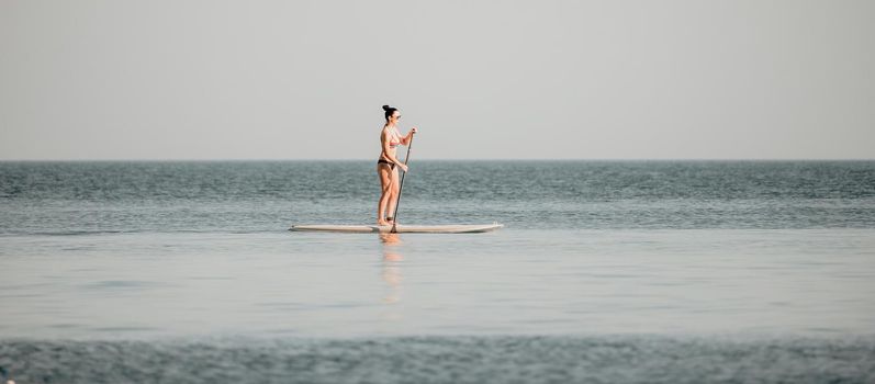 Silhouette of woman standing, surfing on SUP board, confident paddling through water surface. Idyllic sunset or sunrise. Sports active lifestyle at sea or river.