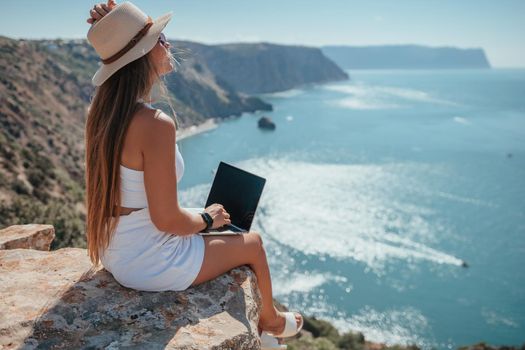 Successful business woman in yellow hat working on laptop by the sea. Pretty lady typing on computer at summer day outdoors. Freelance, travel and holidays concept.