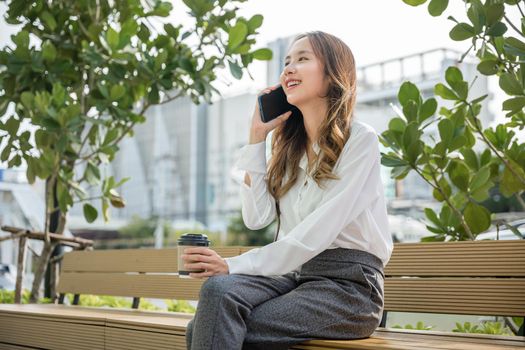 Happy smiling businesswoman sitting talk on mobile phone outside street city near office, Asian business young woman using mobile smartphone talking on cellphone outdoor corporate building exterior