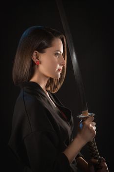 Portrait in profile of a young woman who holds a katana in her hands in front of her on a black background.