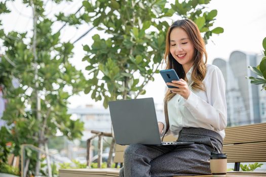 Happy smiling businesswoman sit writing text on mobile phone outside street city near office in morning, Asian business young woman working laptop and using mobile smartphone outdoor building exterior