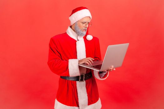 Serious elderly man with gray beard wearing santa claus costume working on computer, looking at display with concentrated facial expression. Indoor studio shot isolated on red background.