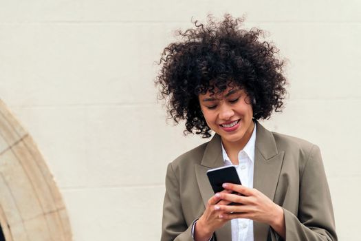 young woman with curly hair smiles happy sending a message with her mobile phone, concept of youth and technology, copy space for text