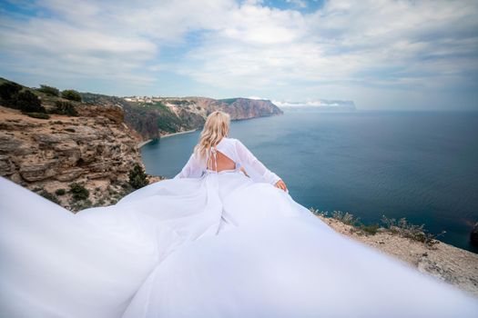 Blonde with long hair on a sunny seashore in a white flowing dress, rear view, silk fabric waving in the wind. Against the backdrop of the blue sky and mountains on the seashore