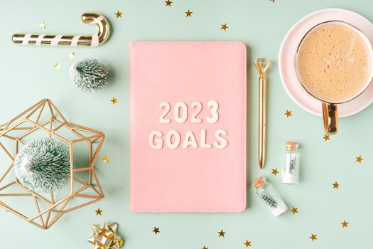 Christmas pink notepad with 2023 goals letters text. Flat lay on green mint table background with planner, cup of coffee, candle, Christmas decoration, notebook and stationery. Top view Desktop.