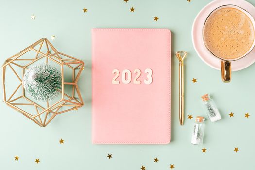 Christmas pink notepad with 2023 letters text. Flat lay on green mint table background with planner, cup of coffee, candle, Christmas decoration, notebook and stationery. Top view Desktop