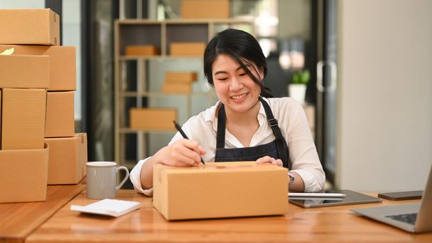 Smiling woman business owner writing address on cardboard box and preparing for shipping. E-commerce, Online selling concept.