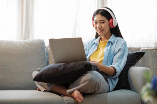 Young asian woman smiling listening to music using laptop surfing the internet while sitting on couch at home. Online connection and online learning concept.