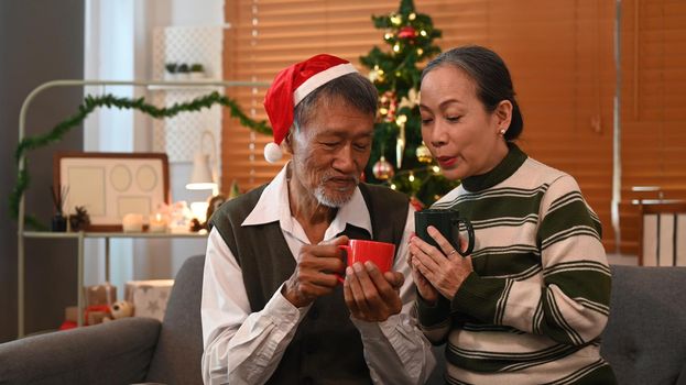 Happy senior couple sitting on couch next to a decorated Christmas tree and enjoying drinking hot chocolate together.