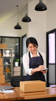 Young woman in apron checking product purchase order, working at home office. E-Commerce, online business, online sales concept.