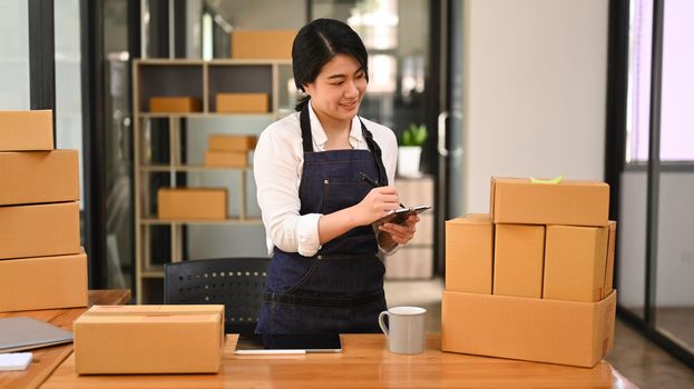 Smiling startup small business entrepreneur checking order and preparing parcel boxes for shipping to customer.